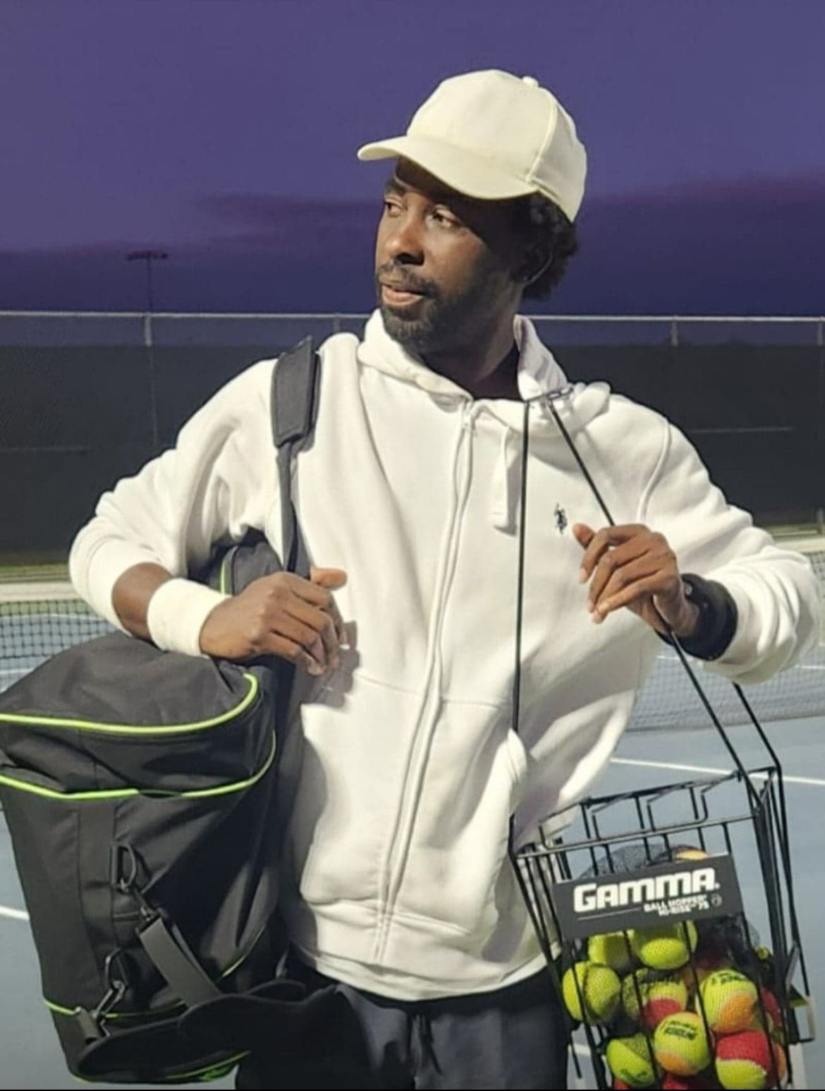 Divine Oshideko at a tennis court carrying his racquet bag and basket of tennis balls